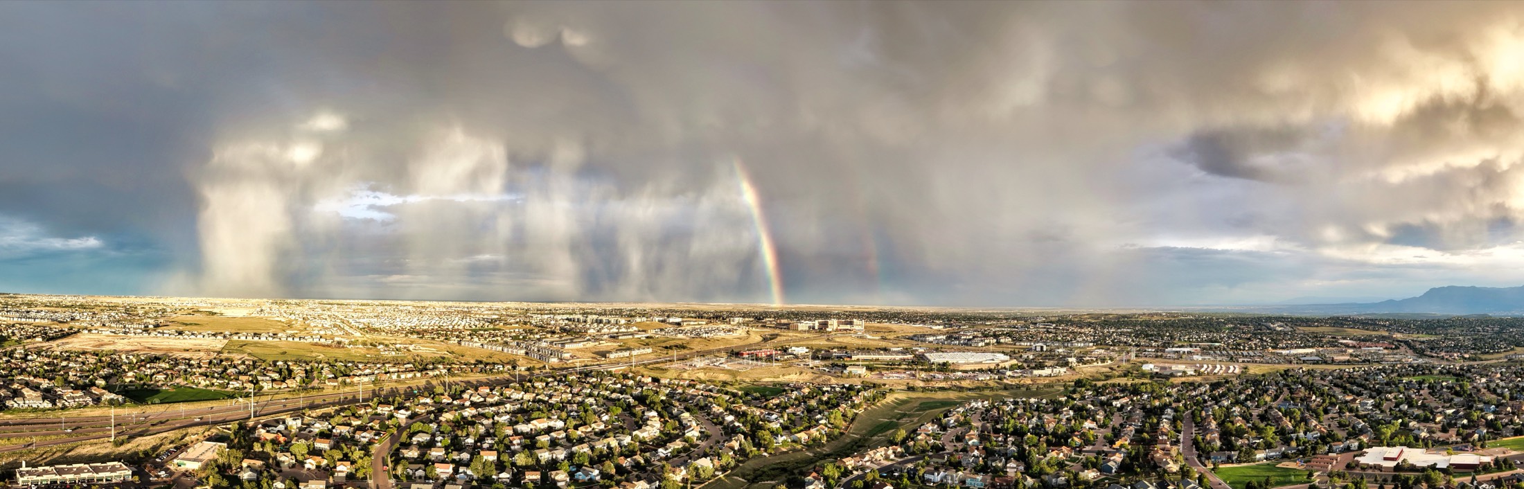 Ultra-wide aerial panorama of a rain squall with a double rainbow breaking over suburban rooftops on the plains